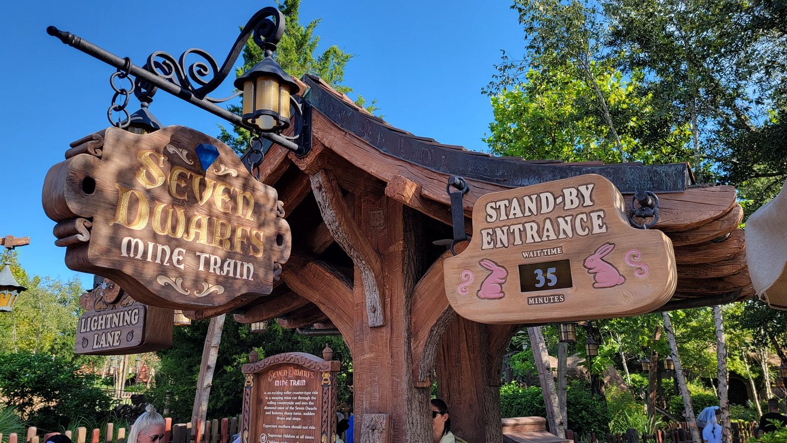 Wooden sign for the Seven Dwarfs Mine Train ride with a Stand by Entrance indicator and rustic lanterns under a blue sky