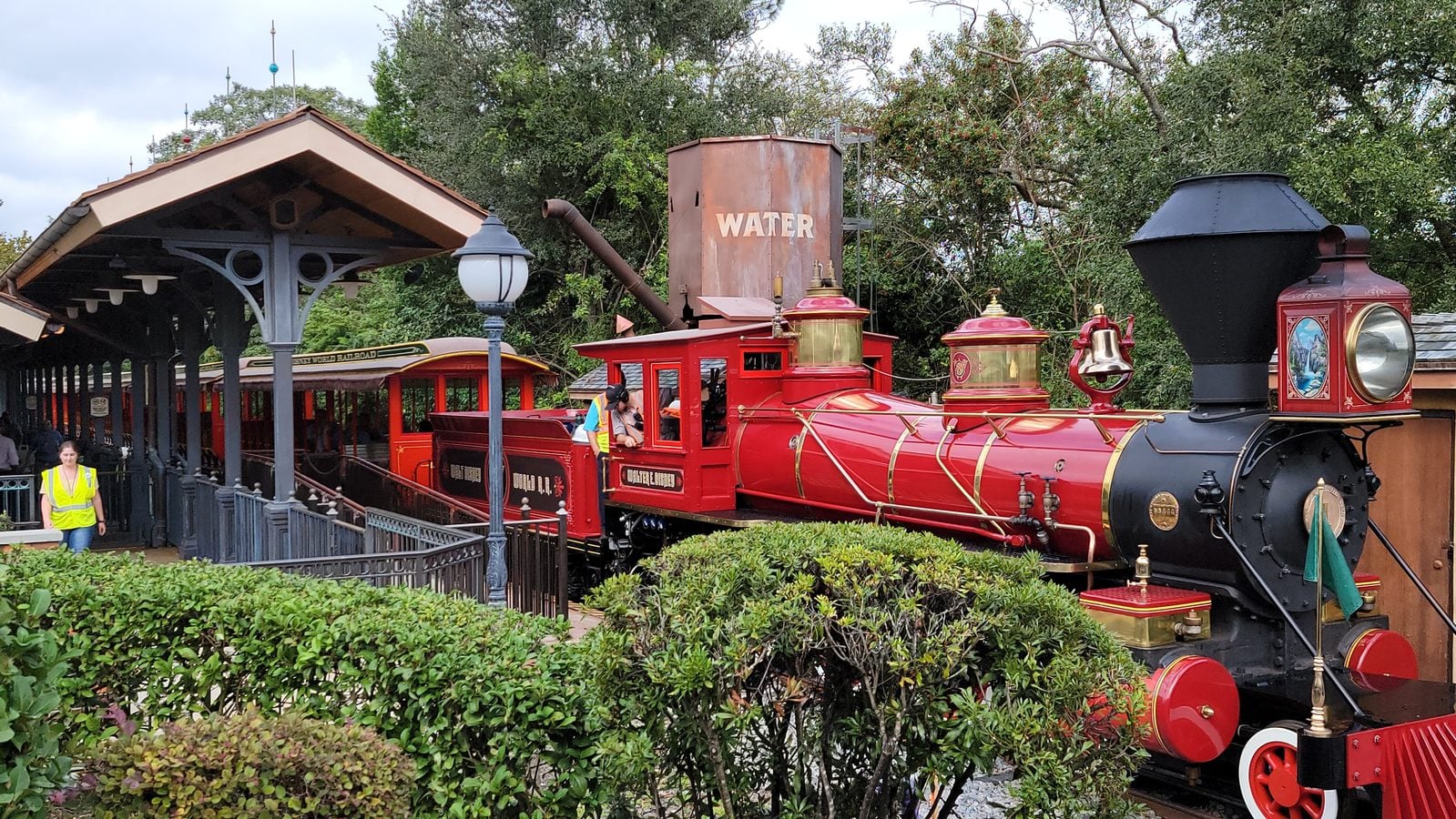 Vintage red steam locomotive at a station platform with a sheltered waiting area and crew member in a high visibility vest nearby