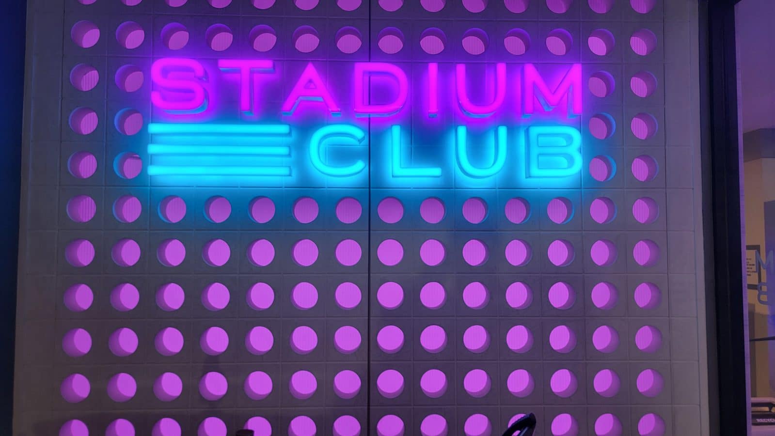 Pink and blue neon sign reading STADIUM CLUB on a wall with purple circular panels behind it
