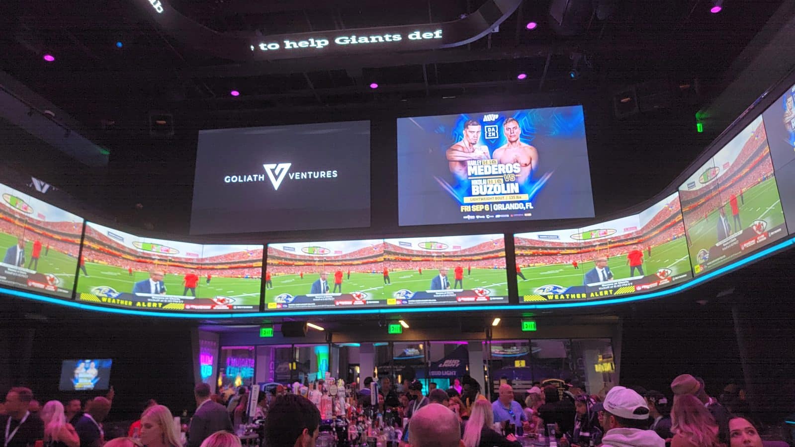 Indoor event venue with curved video walls showing a boxing poster Goliath Ventures logo and spectators at tables beneath