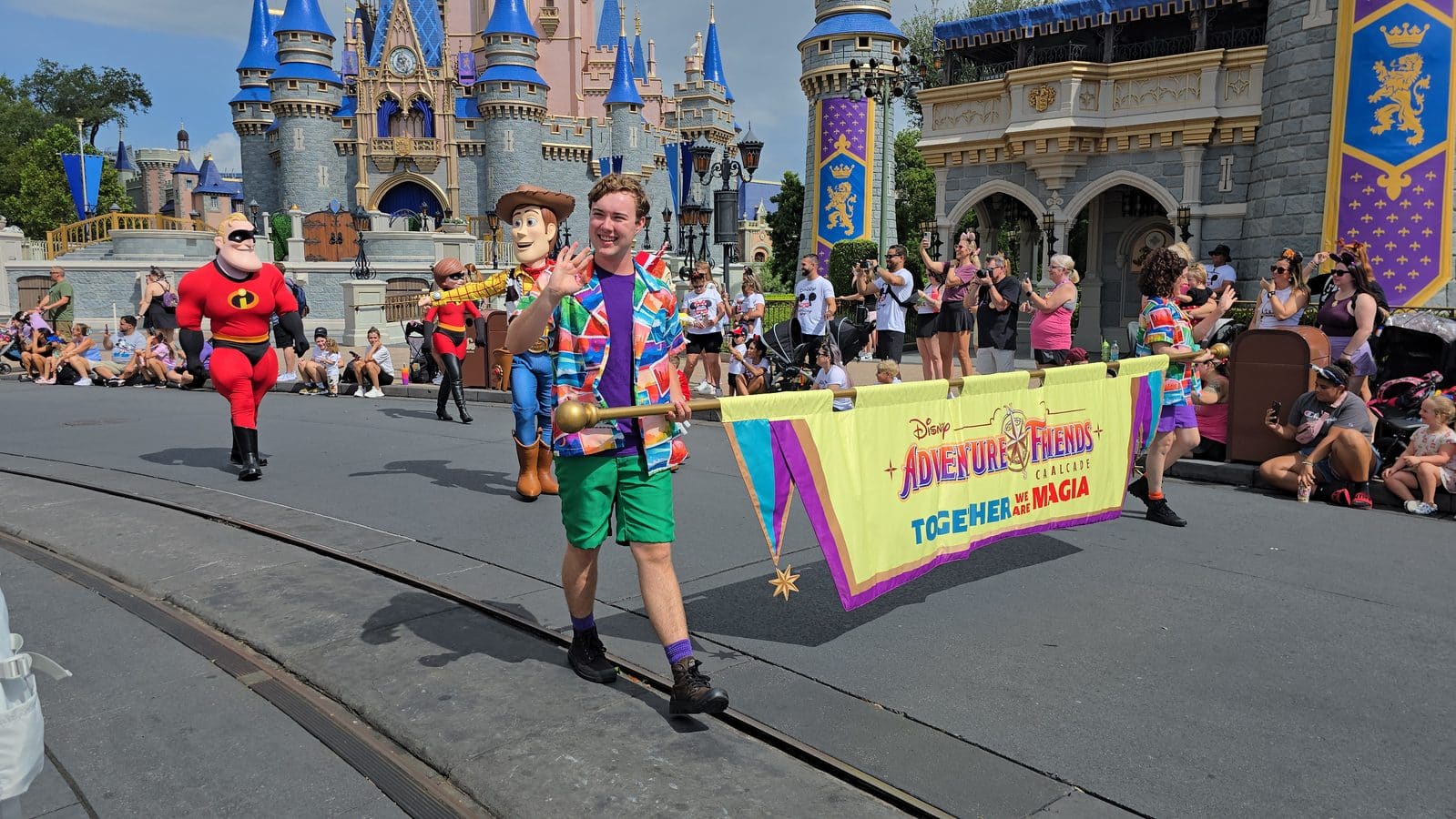 Parade scene at a Disney park a man in a bright multicolored shirt waves to cheering crowds flank​ed by costumed characters and a rainbow banner that reads Adventure Friends Cavalcade a castle backdrop