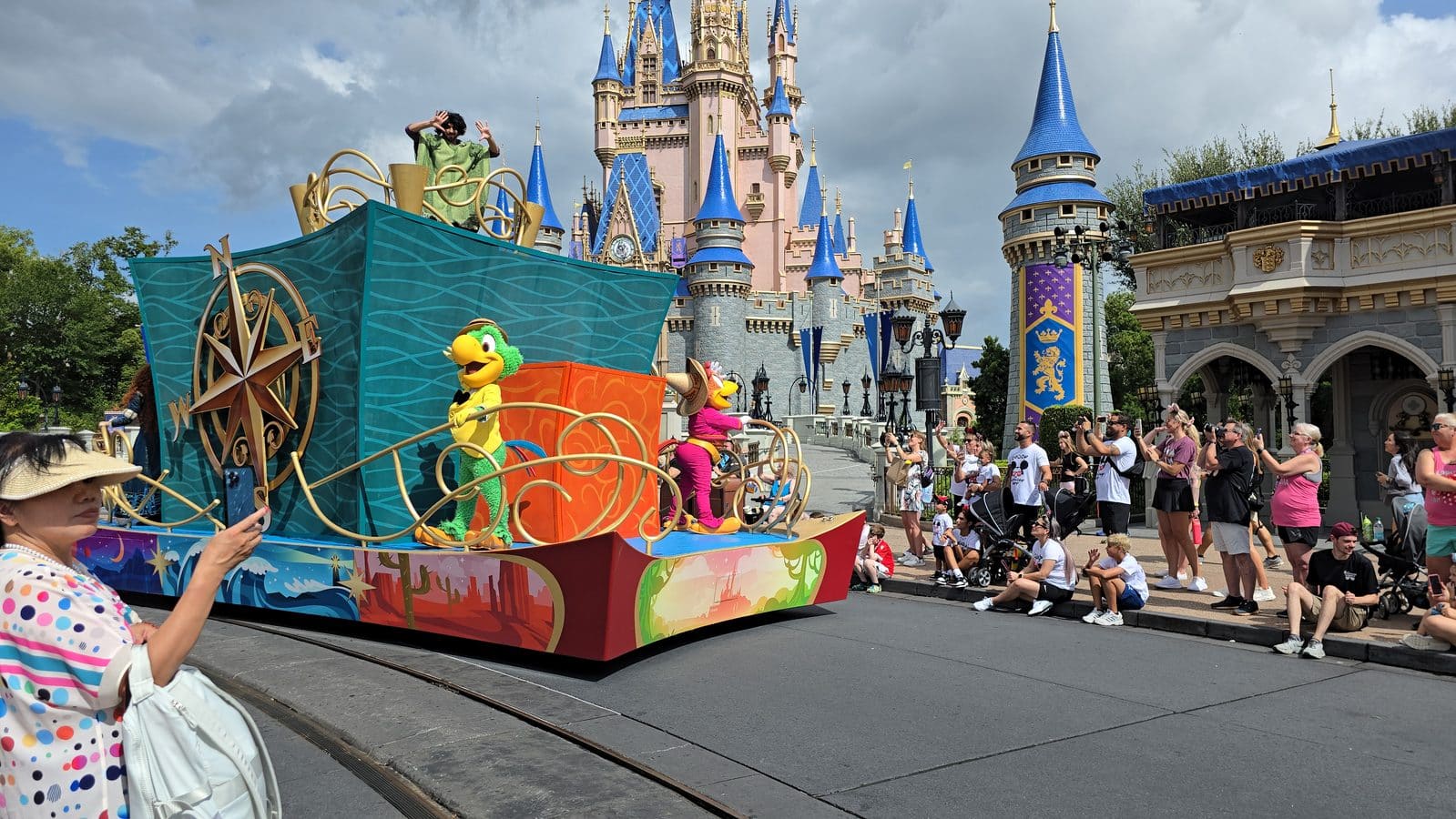 Colorful parade float with cartoon bird performers in front of a pink and blue castle crowd watching along a theme park street