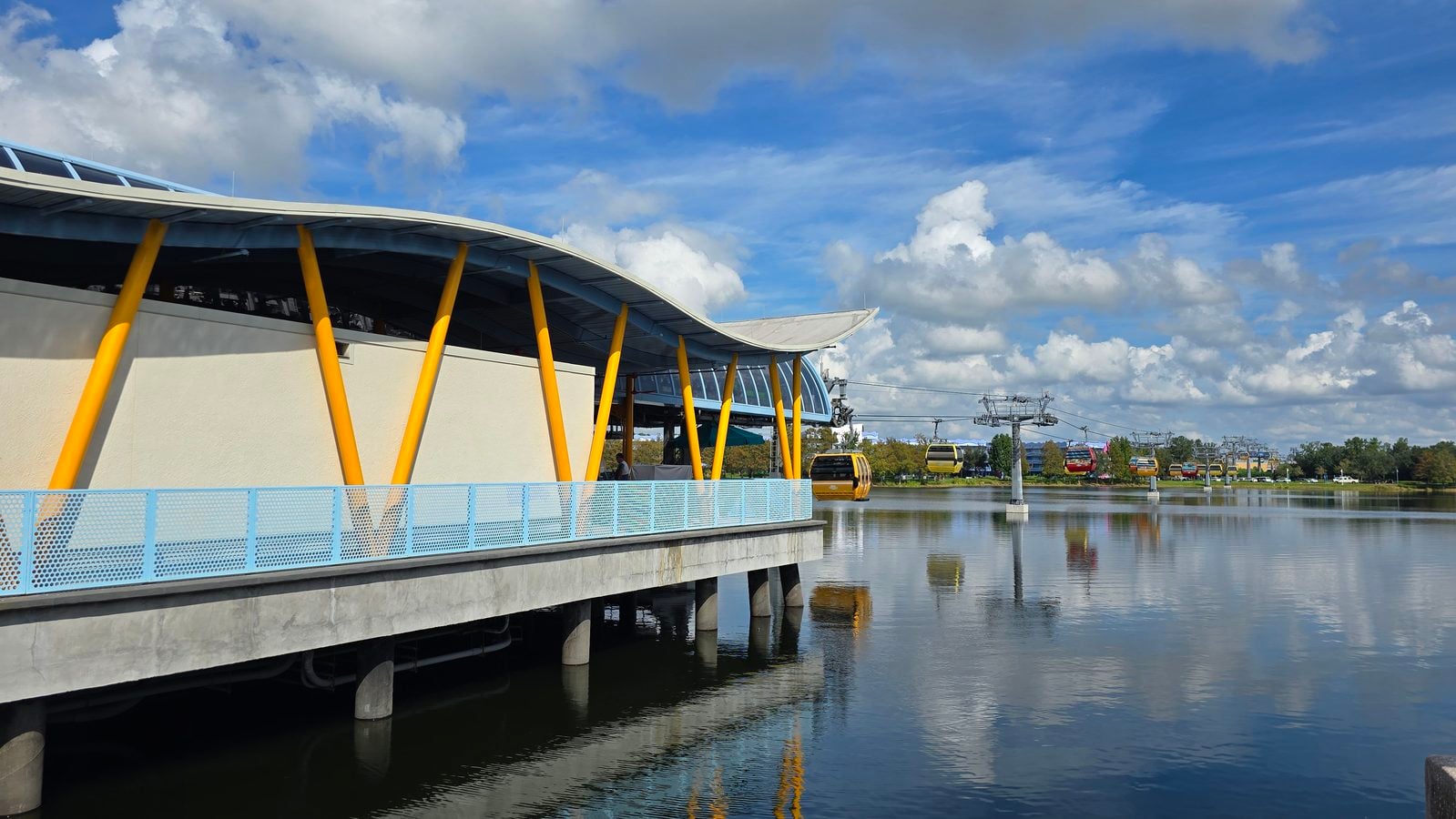 Modern waterfront building with bright yellow supports and light blue railing overlooking a calm lake gondola ride in the background under a blue sky with clouds