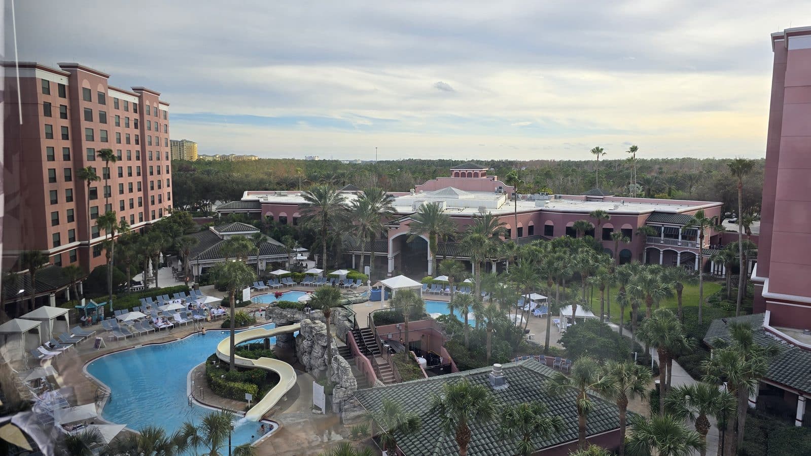 Aerial view of a pink hued resort with multiple buildings palm trees and a winding pool deck below under a cloudy sky