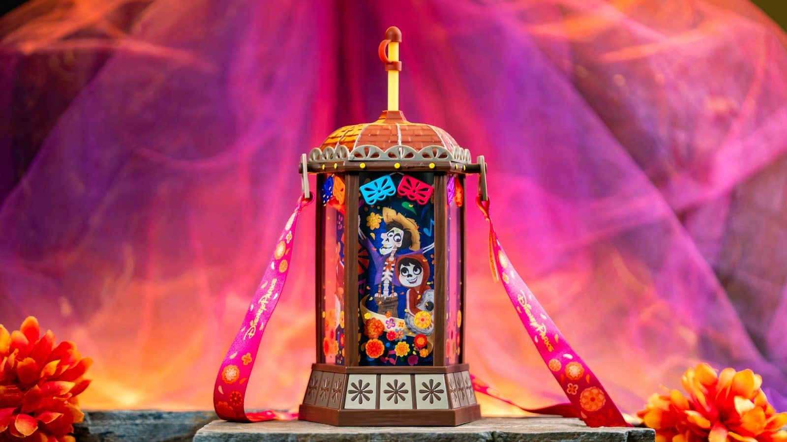 Colorful decorative lantern with Day of the Dead motifs and pink ribbons set against a vibrant pink orange background