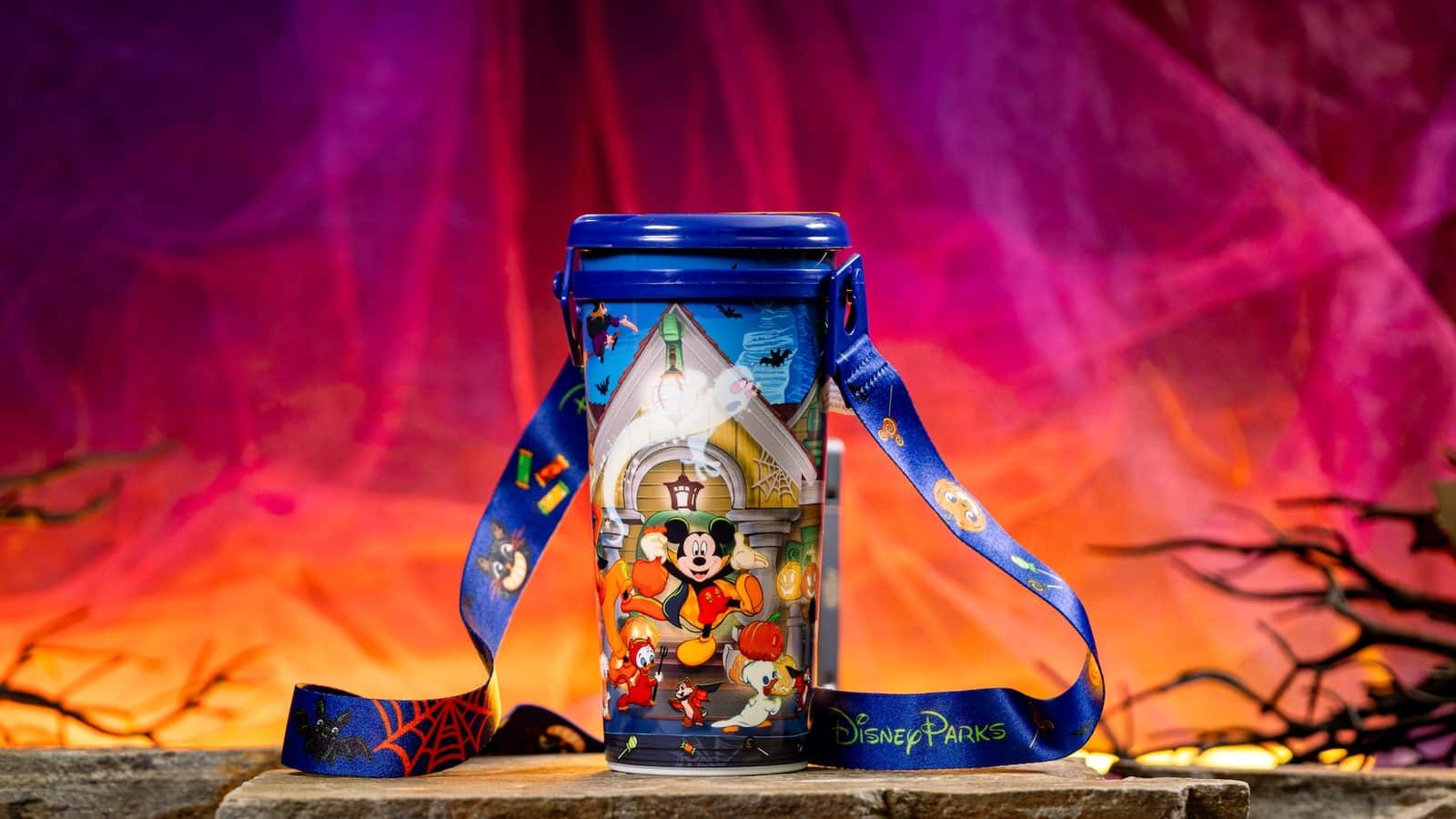 Blue Disney Parks cup with Halloween designs and a blue lid resting on a wooden surface against a pinkorange background with a ribbon