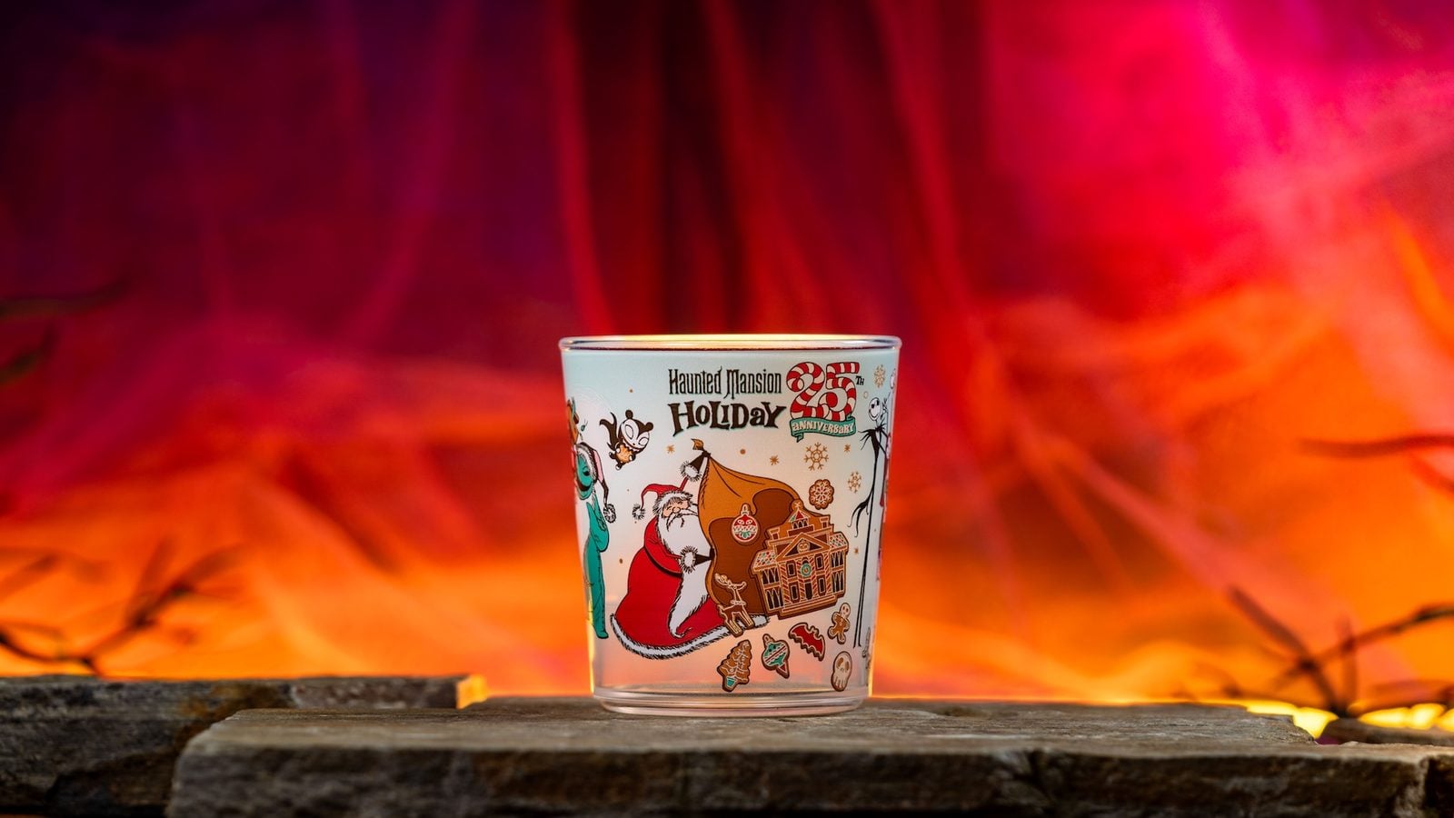 Cup featuring Haunted Mansion Holiday artwork with Santa a gingerbread house and cartoon characters against an orangepink fiery background on a wooden surface