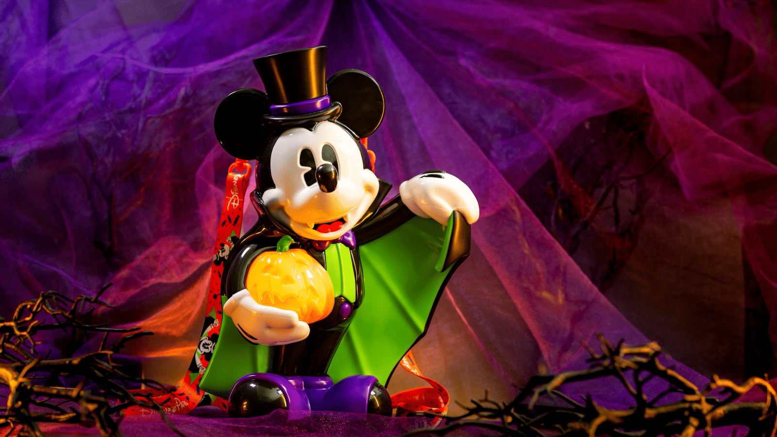 Mickey Mouse in a green cape and purple bow tie holds a glowing jack o lantern against a purple fabric backdrop wearing a top hat for Halloween fun