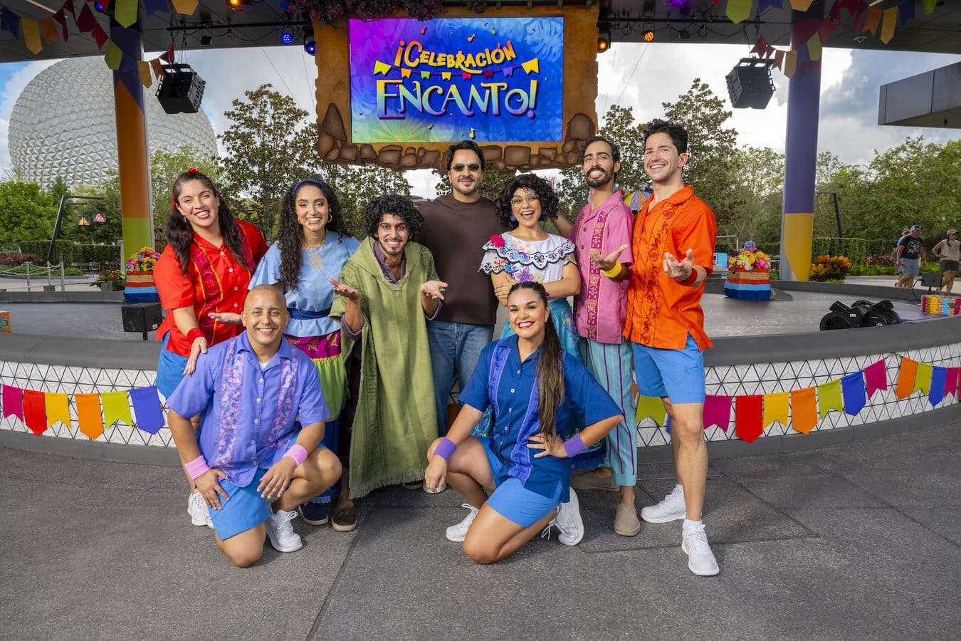 Group of performers in colorful outfits posing on a stage with a bright screen reading Celebración Encanto behind them