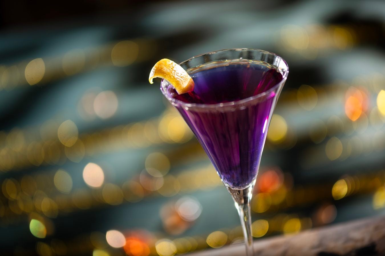 Purple cocktail in a martini glass with an orange peel garnish against blurred bar lights