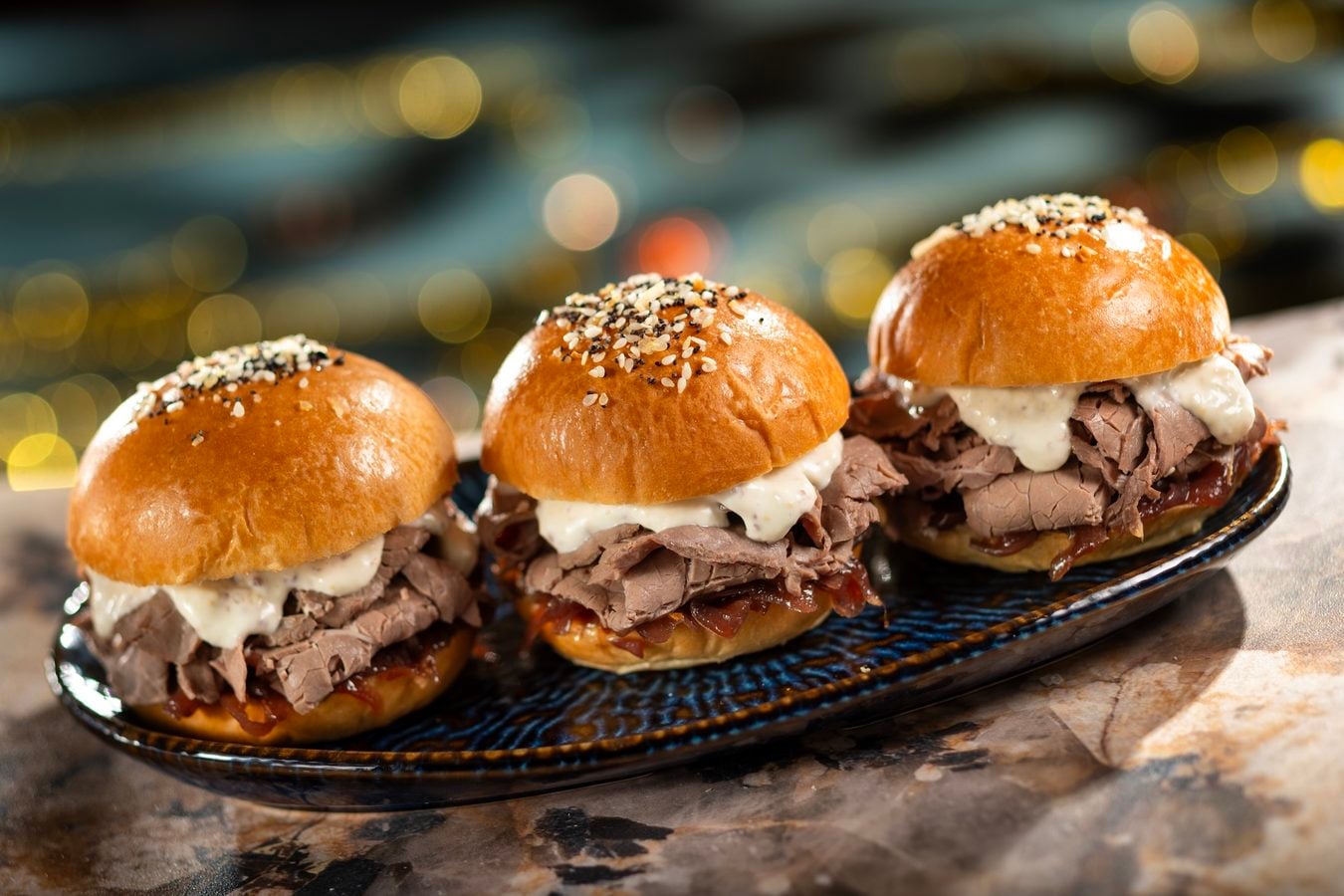 Three beef sliders with sesame buns and creamy white sauce on a dark plate festive bokeh background
