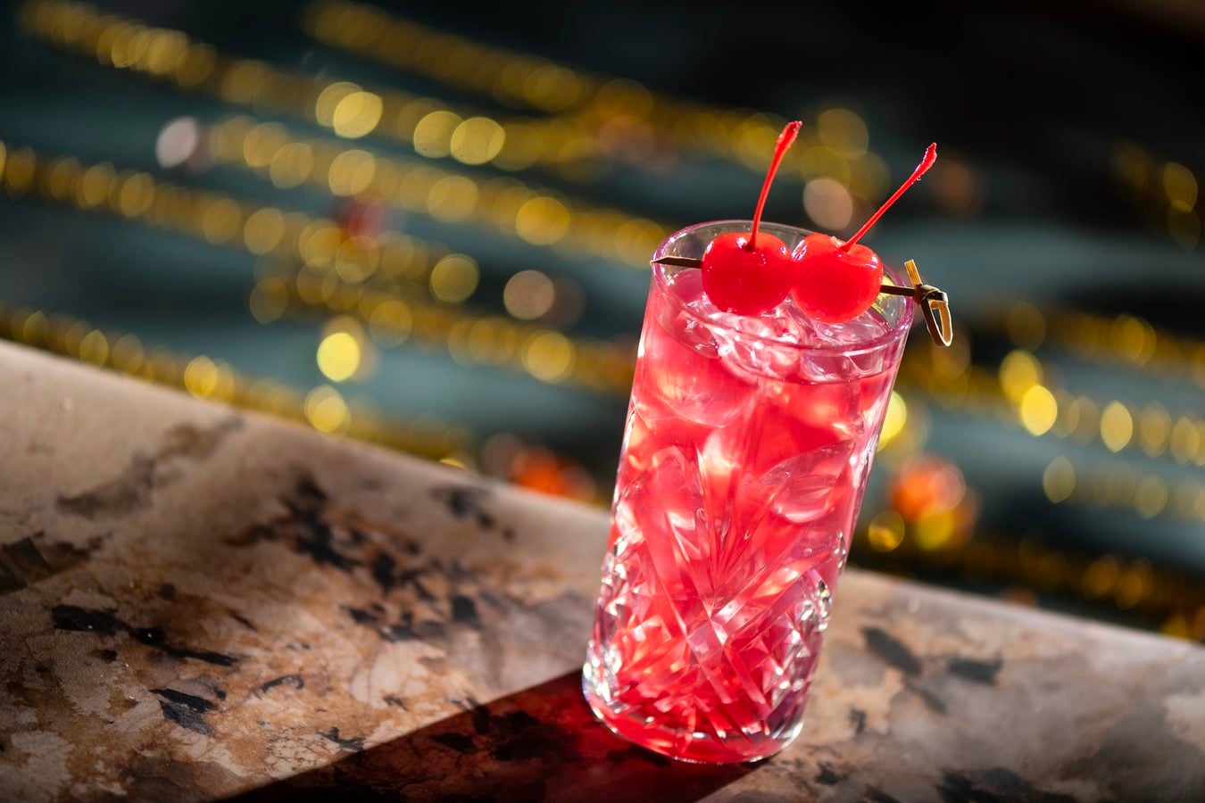 A pink cocktail with two cherries on a skewers in a crystal glass on a bar with blurred string lights in the background