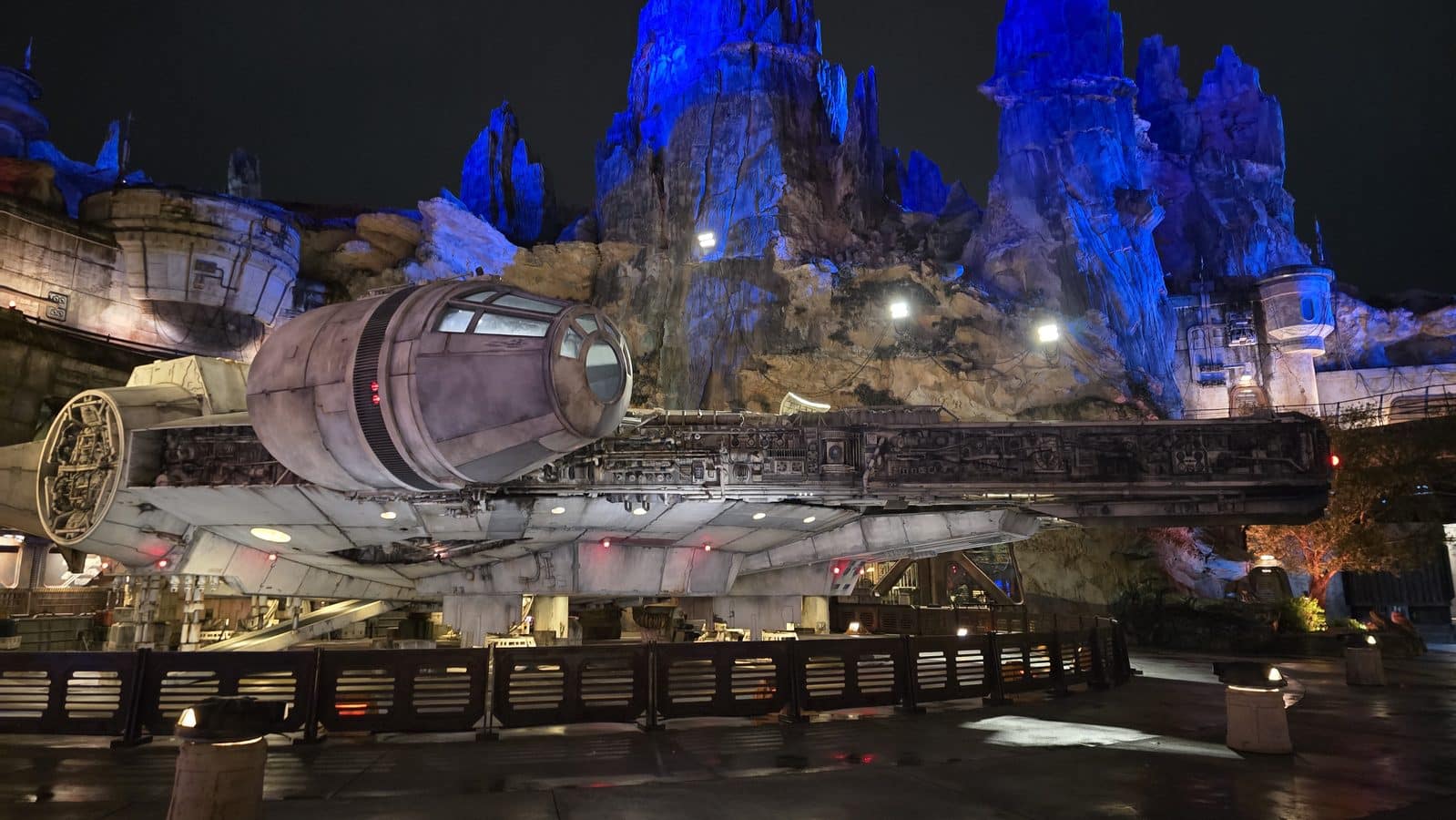 Nighttime view of a large Millennium Falcon spaceship replica on a track with blue lit rocky cliffs in the background and a low barrier in the foreground