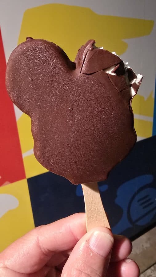 Chocolate coated ice cream bar on a wooden stick with a bite taken out against a colorful background