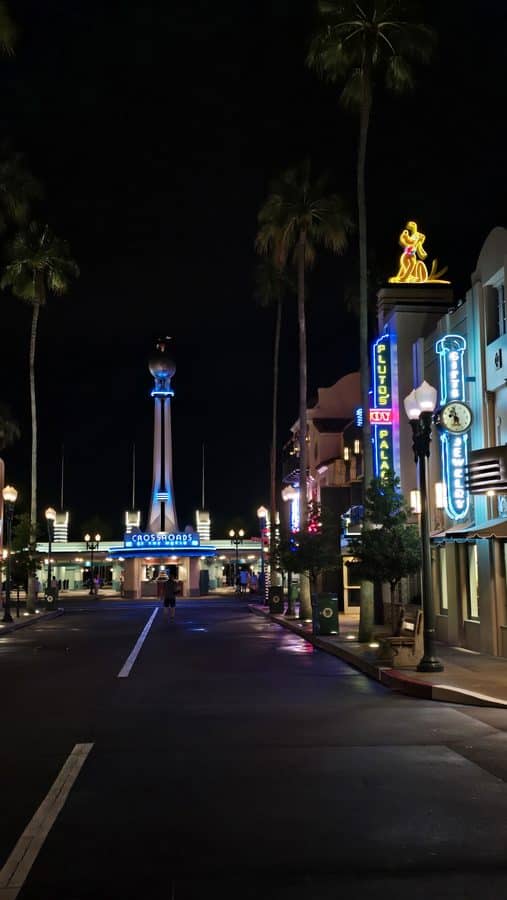 Nighttime street scene with palm trees neon shop signs and a tall illuminated tower at a Crossroads entrance