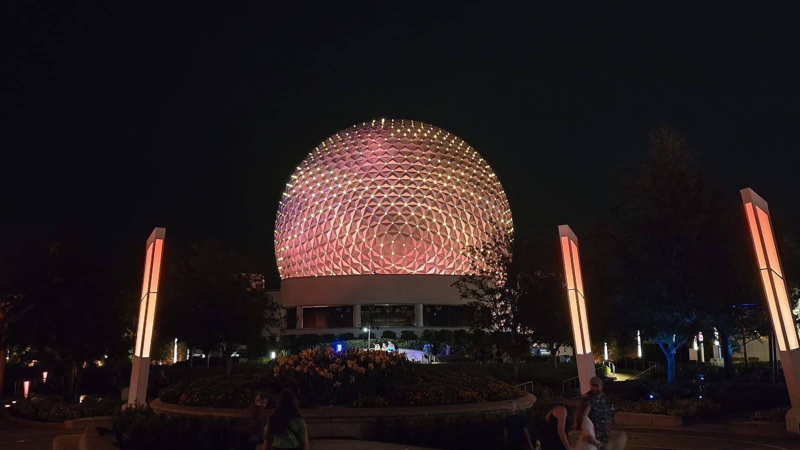 Night view of a pink illuminated geodesic dome surrounded by tall orange lit pillars at night in a park setting