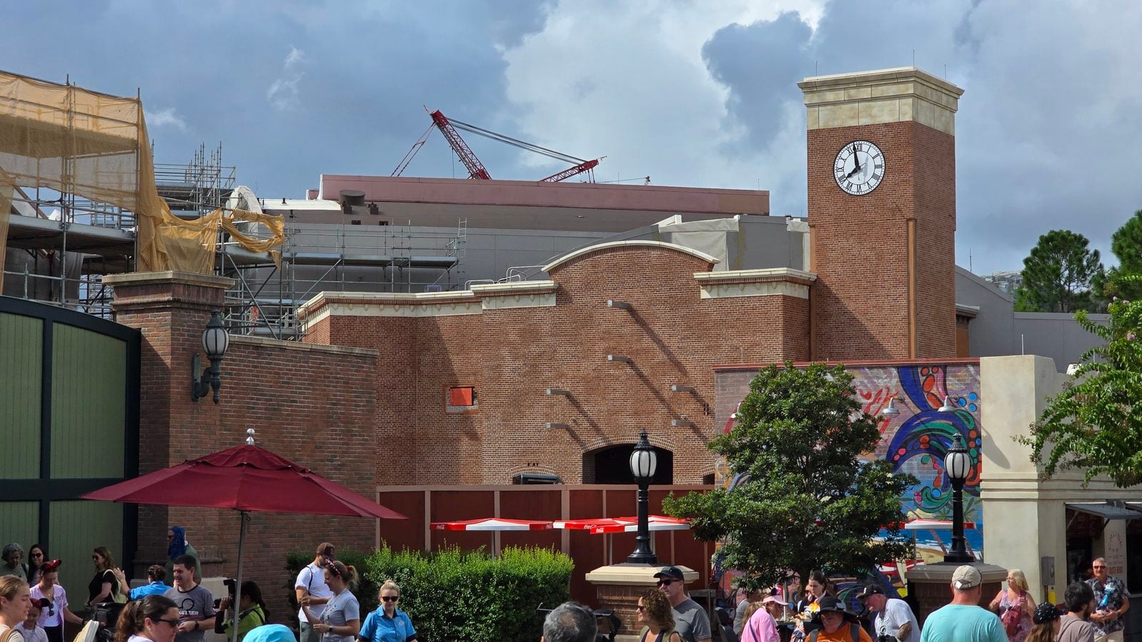 Brick clock tower building with construction scaffolding on the left people gathered in a sunny outdoor plaza with red umbrellas