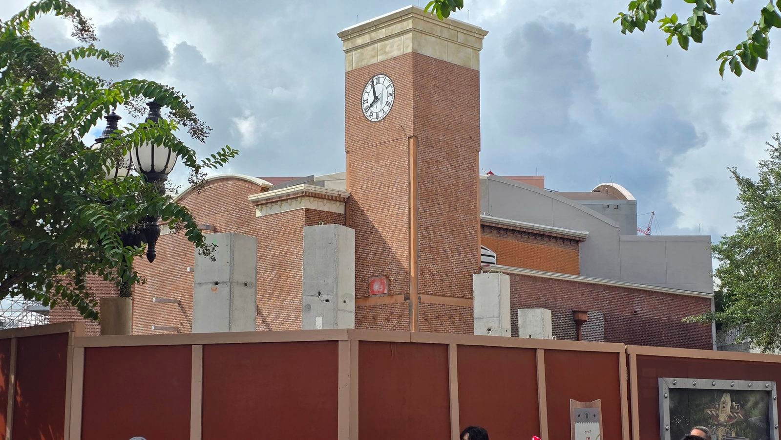 Brick clock tower rising from a red brick building framed by a brown construction fence and green trees against a cloudy sky