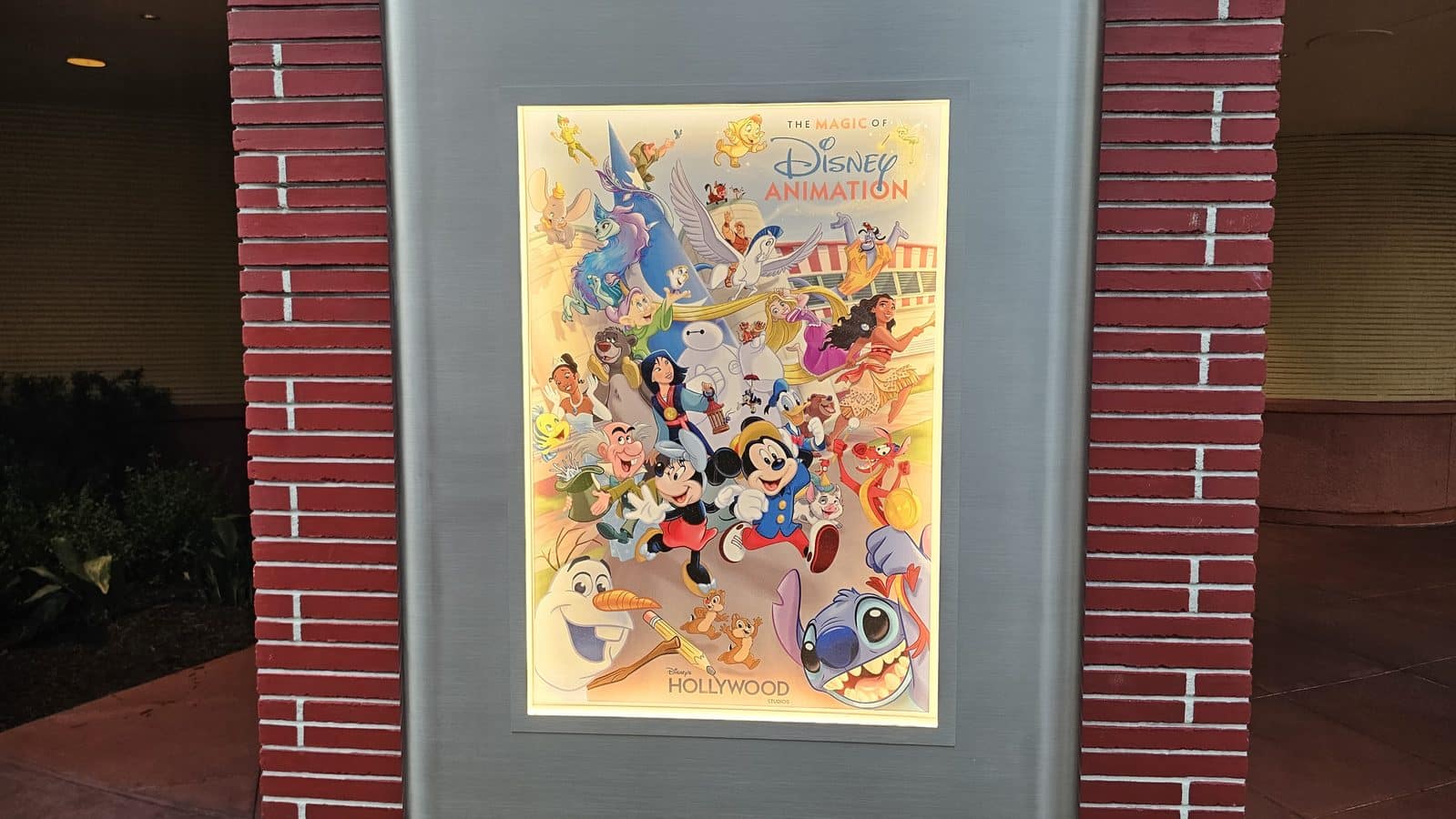 Colorful Disney Animation poster framed on a gray pedestal set against a red brick wall outside a building