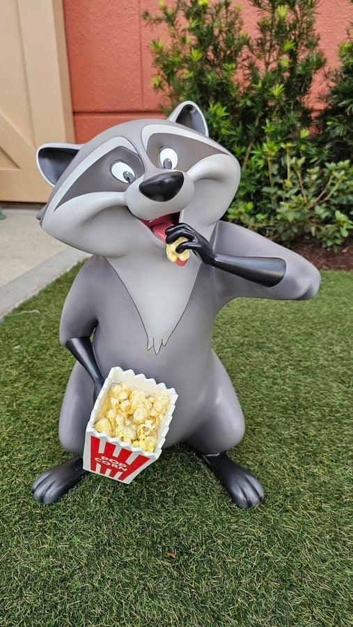 Gray raccoon statue chewing popcorn from a popcorn bucket held in its paw outdoors on a grassy lawn with shrubs in the background