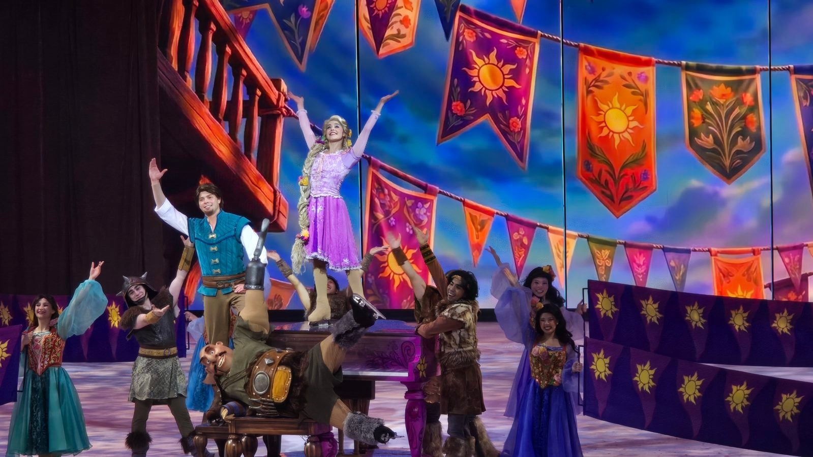 Group of performers in colorful costumes on a stage with sun themed banners central woman in purple dress is raised on a platform while others cheer