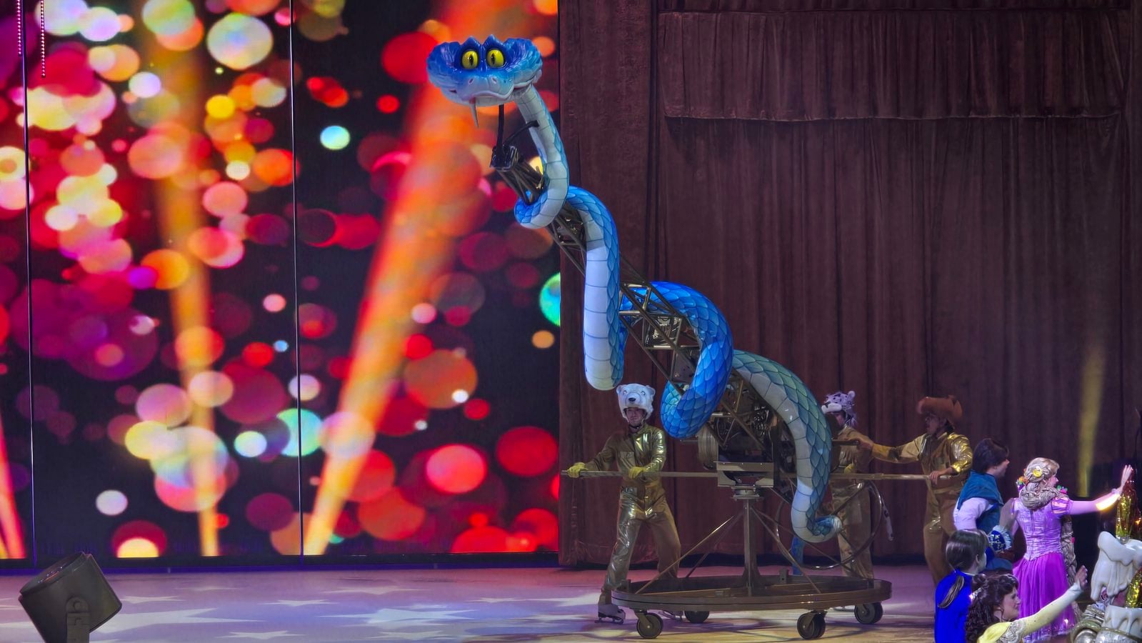 Blue dragon puppet on a wheeled stage prop surrounded by gold costumed performers on a colorful bokeh backdrop stage