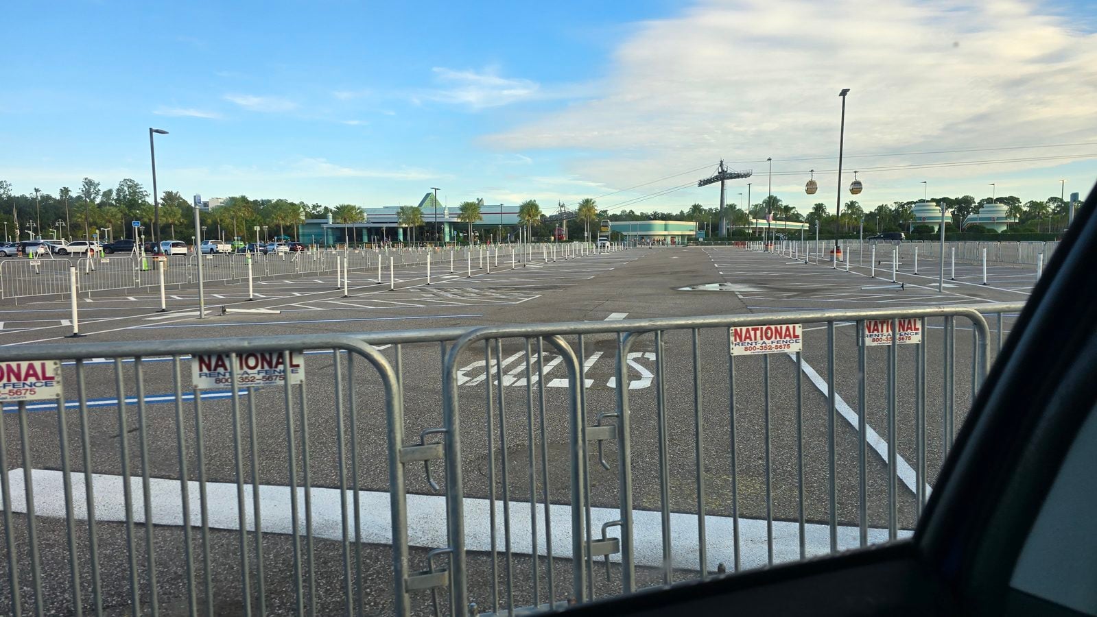 Empty parking lot with white metal barricades in the foreground and a distant building with palm trees under a blue sky informative