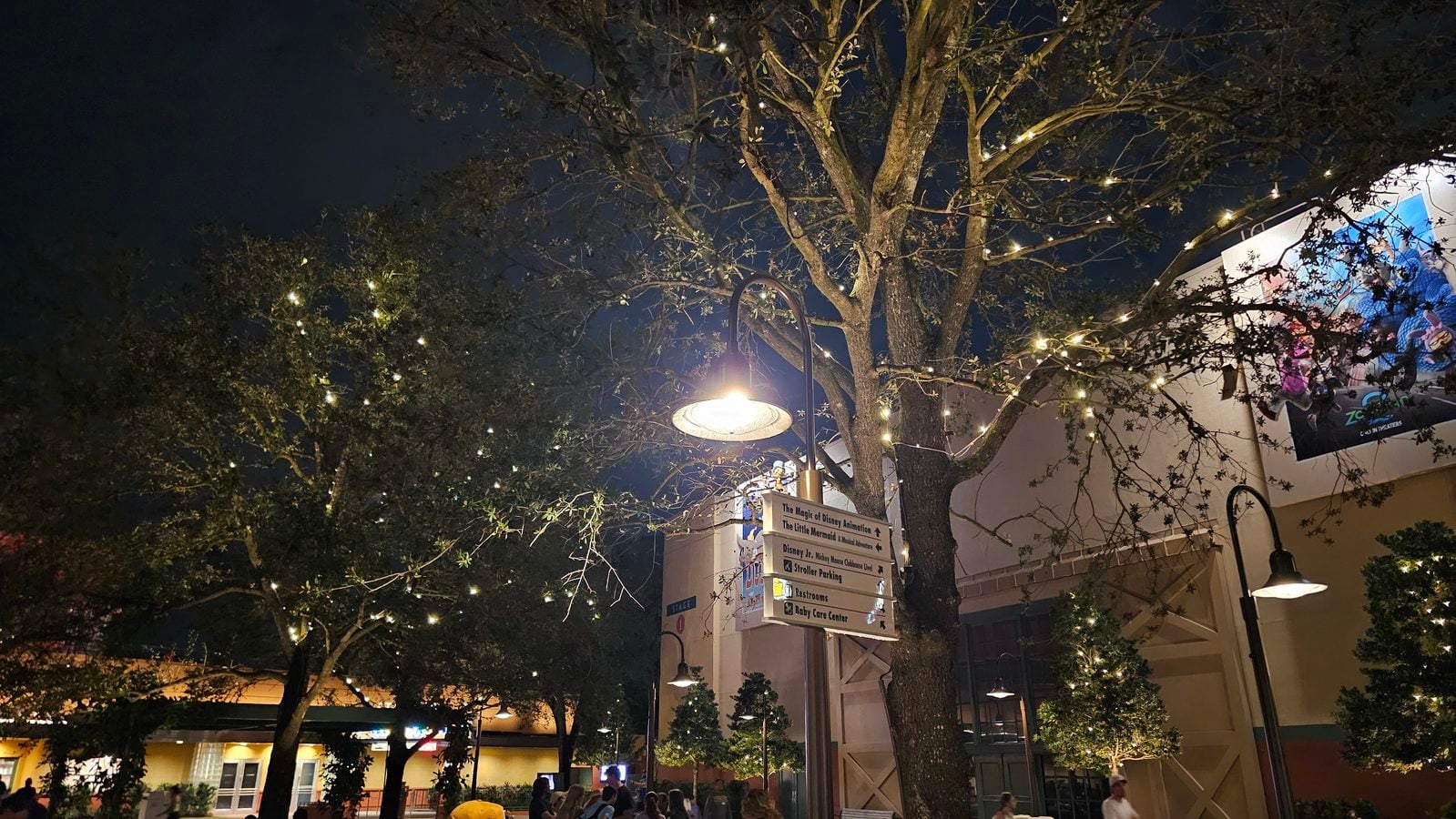 Nighttime outdoor plaza with string lights in tree branches a curved lamppost and people seated near a building with signs