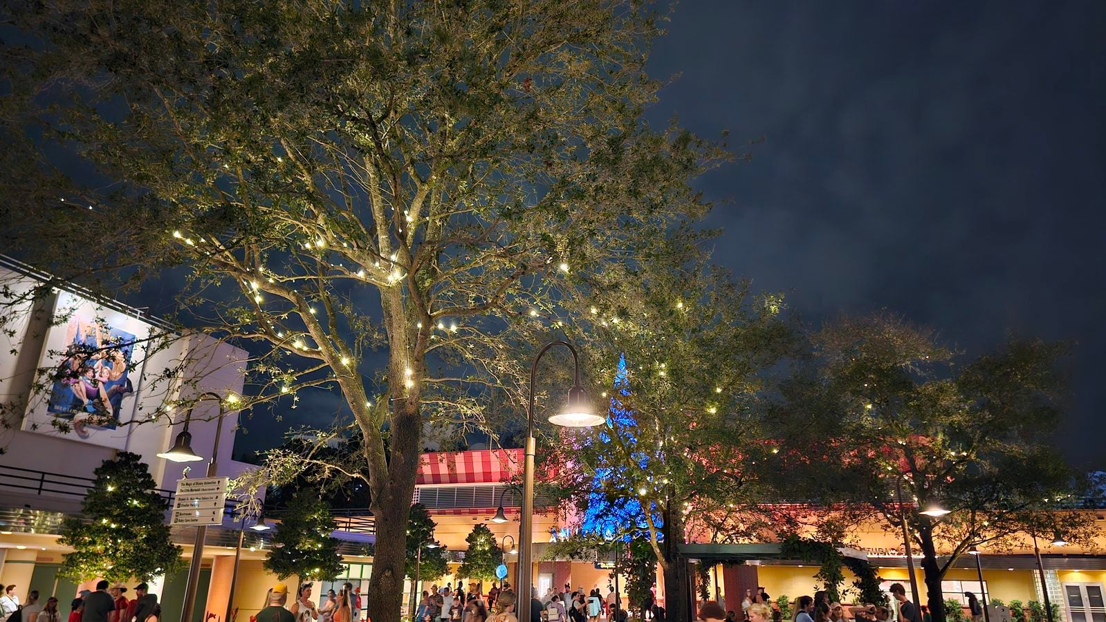 Nighttime outdoor plaza with a large tree wrapped in warm string lights and a crowd of people beneath storefronts in the background