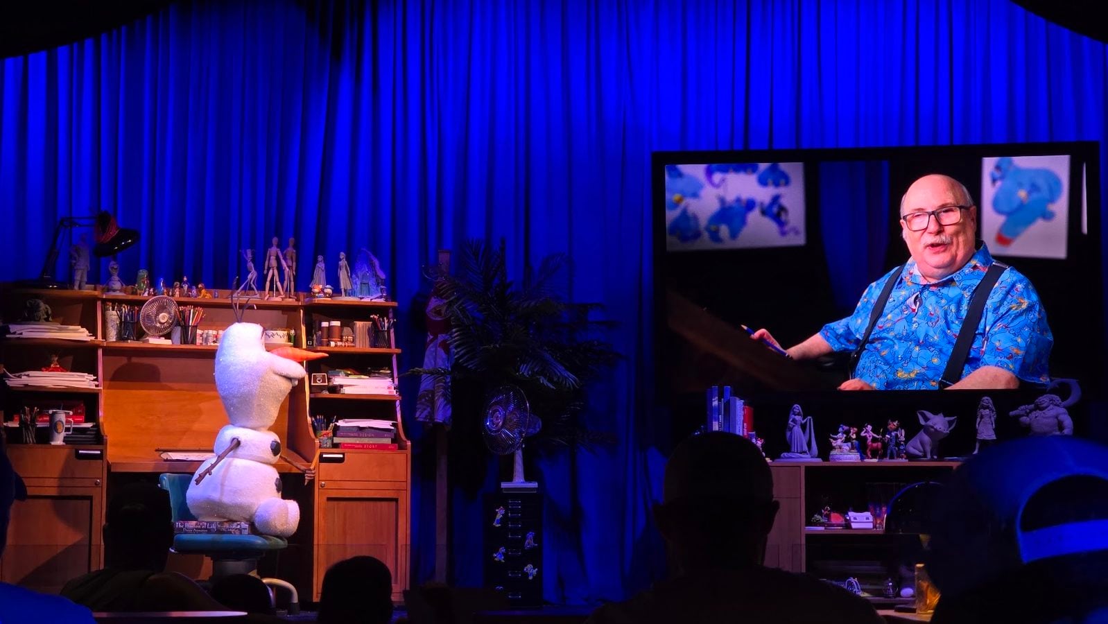 Ventri­loquist on a blue lit stage with a large Snoopy plush on a chair and a cluttered desk of art supplies behind