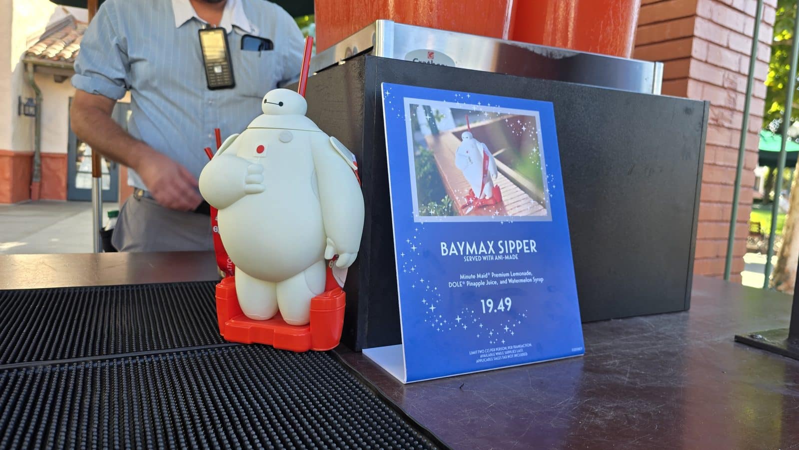 Life size Baymax figure with a red base beside a blue poster reading Baymax Sipper on a kiosk counter on a sunny outdoor setup