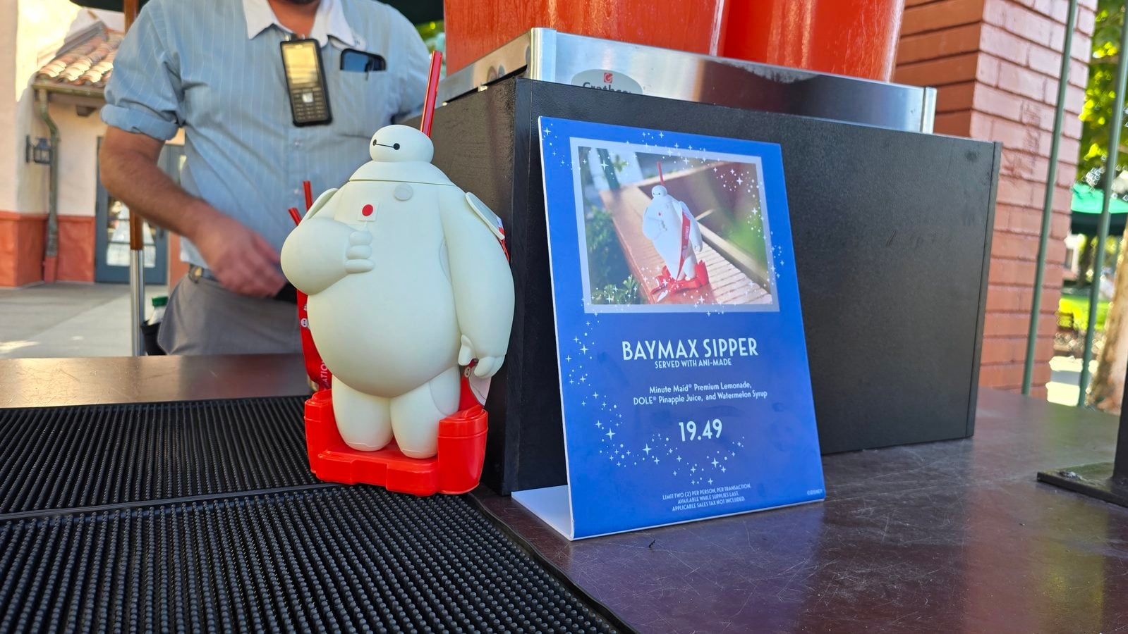 Baymax toy figure with red base beside a blue Baymax Sipper promotion poster on a counter