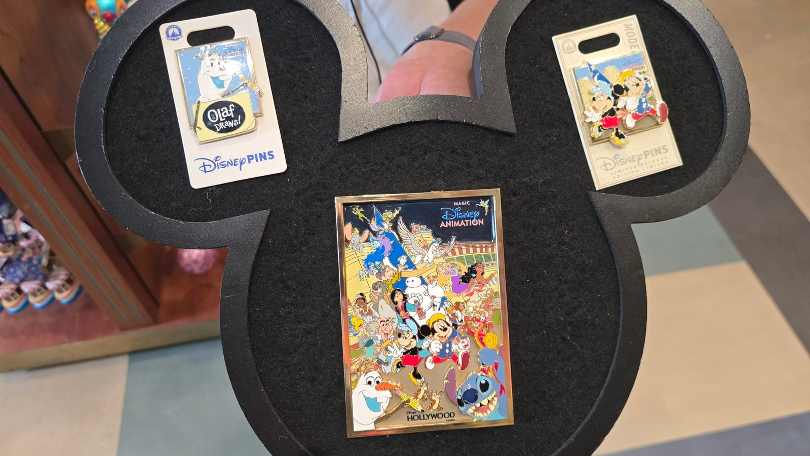Disney pins display on a black circular stand featuring Olaf Draws a Disney Pins card with Mickey and a colorful Disney Animation poster surrounded by characters