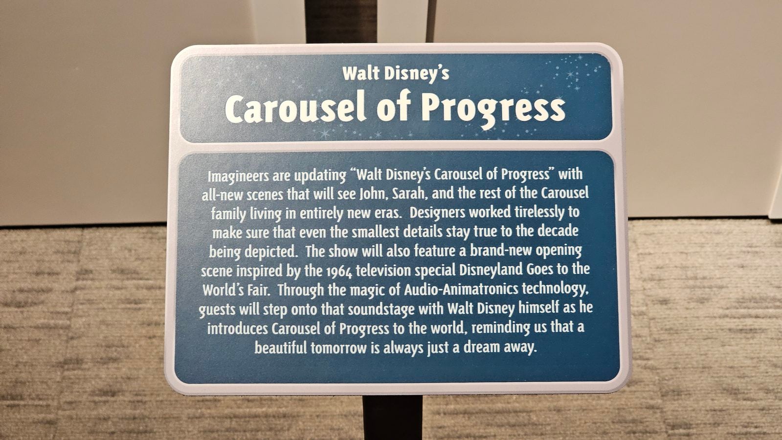 Blue informational sign reading Walt Disneys Carousel of Progress with a paragraph describing the show on a wooden stand edge