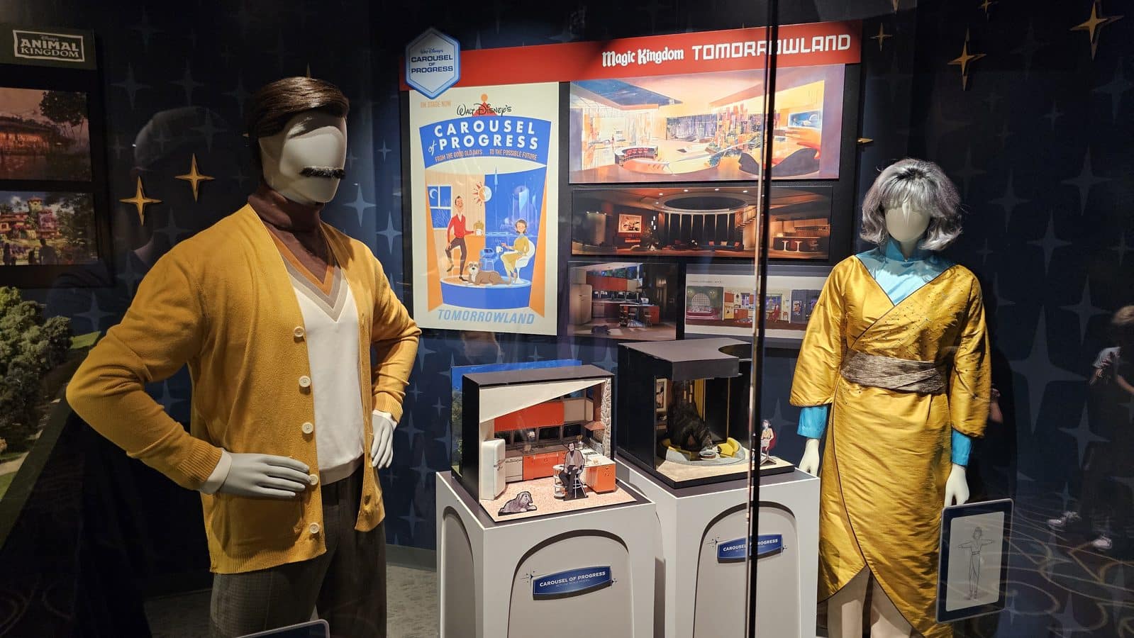 Two life size mannequins in vintage yellow outfits stand in a glass display about Disneys Tomorrowland with Carousel of Progress posters behind them