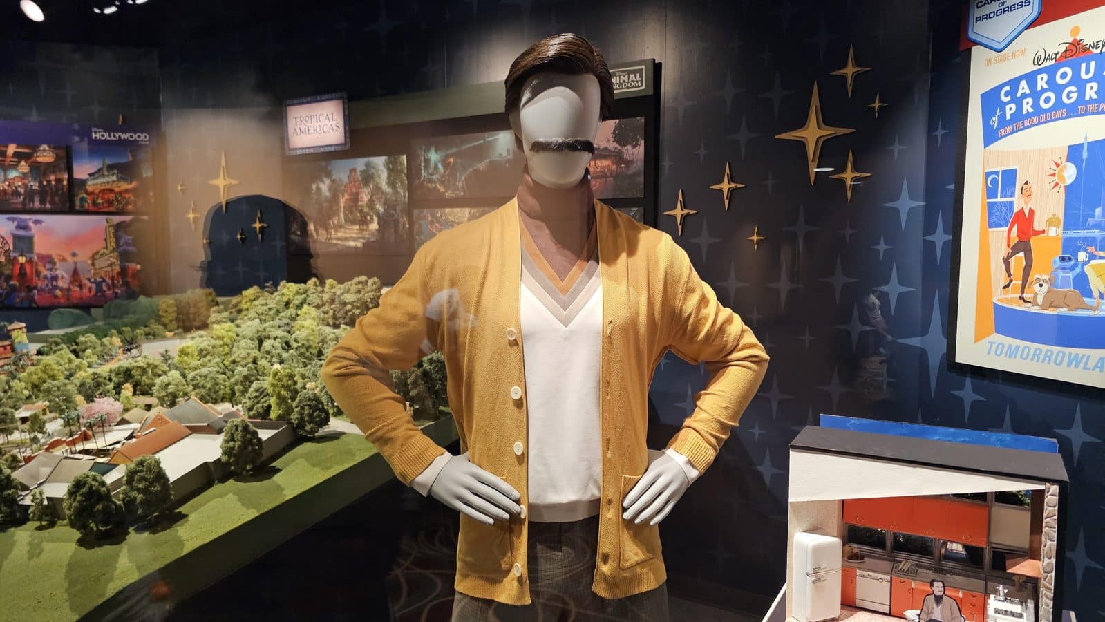 Life size mannequin wearing a mustard cardigan and white V neck shirt in a display window with a miniature landscape to the left and travel posters to the right