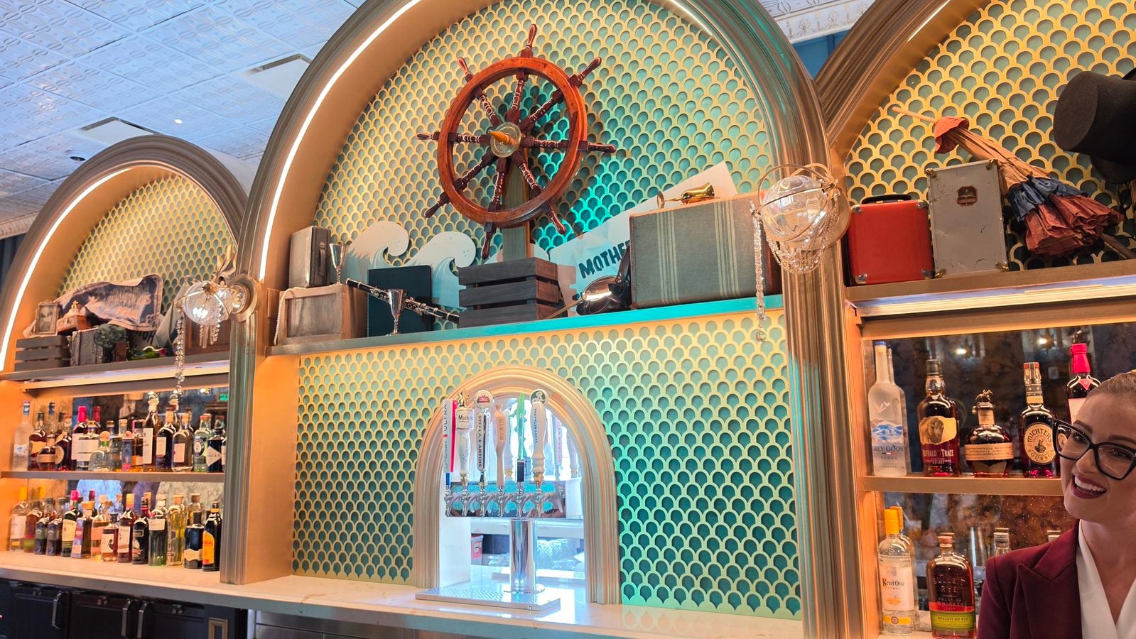 Bar interior with turquoise lattice walls a hanging wooden ship wheel plus bottles on the back bar and a smiling bartender on the right