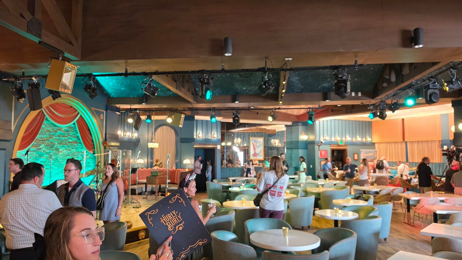 Busy restaurantlounge with turquoise chairs and round tables stage area on the left and colorful lighting overhead and people mingling