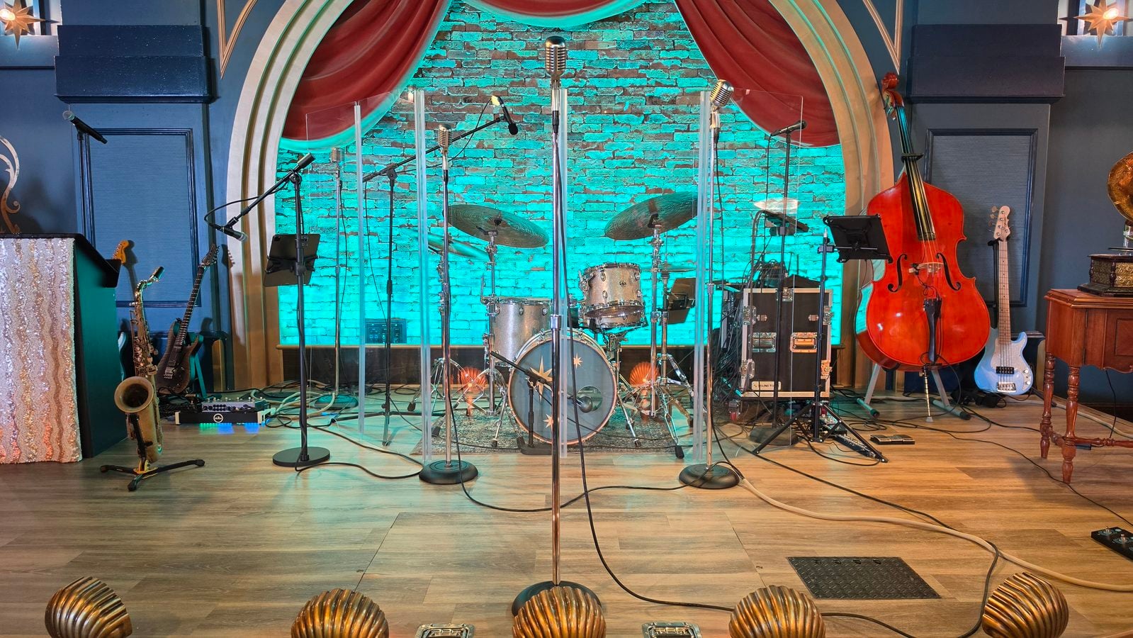 Stage setup for a band drums microphones and a double bass on teal lit brick backdrop with red curtains