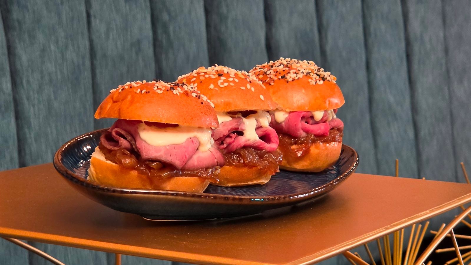 Two beef sliders with melted cheese on sesame buns served on a plate against a blue wooden backdrop
