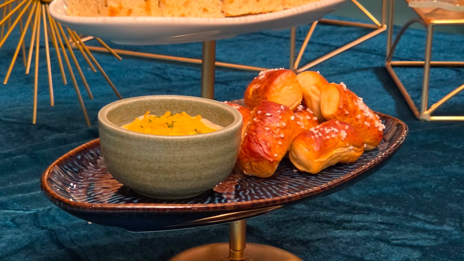 Pretzel knots with coarse salt on a decorative plate beside a green bowl of yellow dip set on a blue tablecloth
