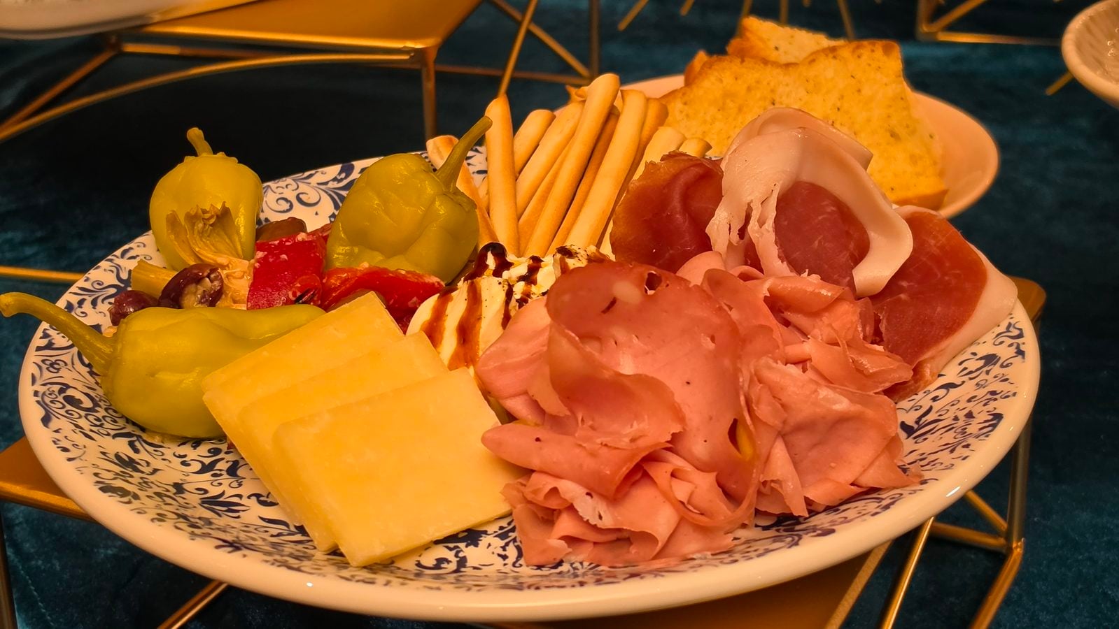 Charcuterie plate with prosciutto sliced cheese olives peppers breadsticks and crackers on a patterned dish