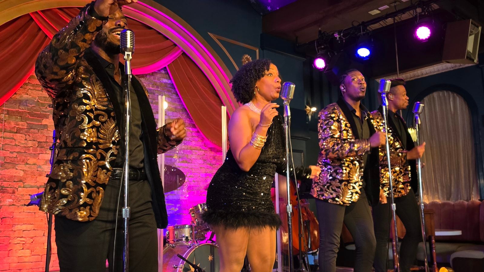 Four singers perform on a brick walled stage with vintage microphones wearing matching gold patterned jackets and black outfits