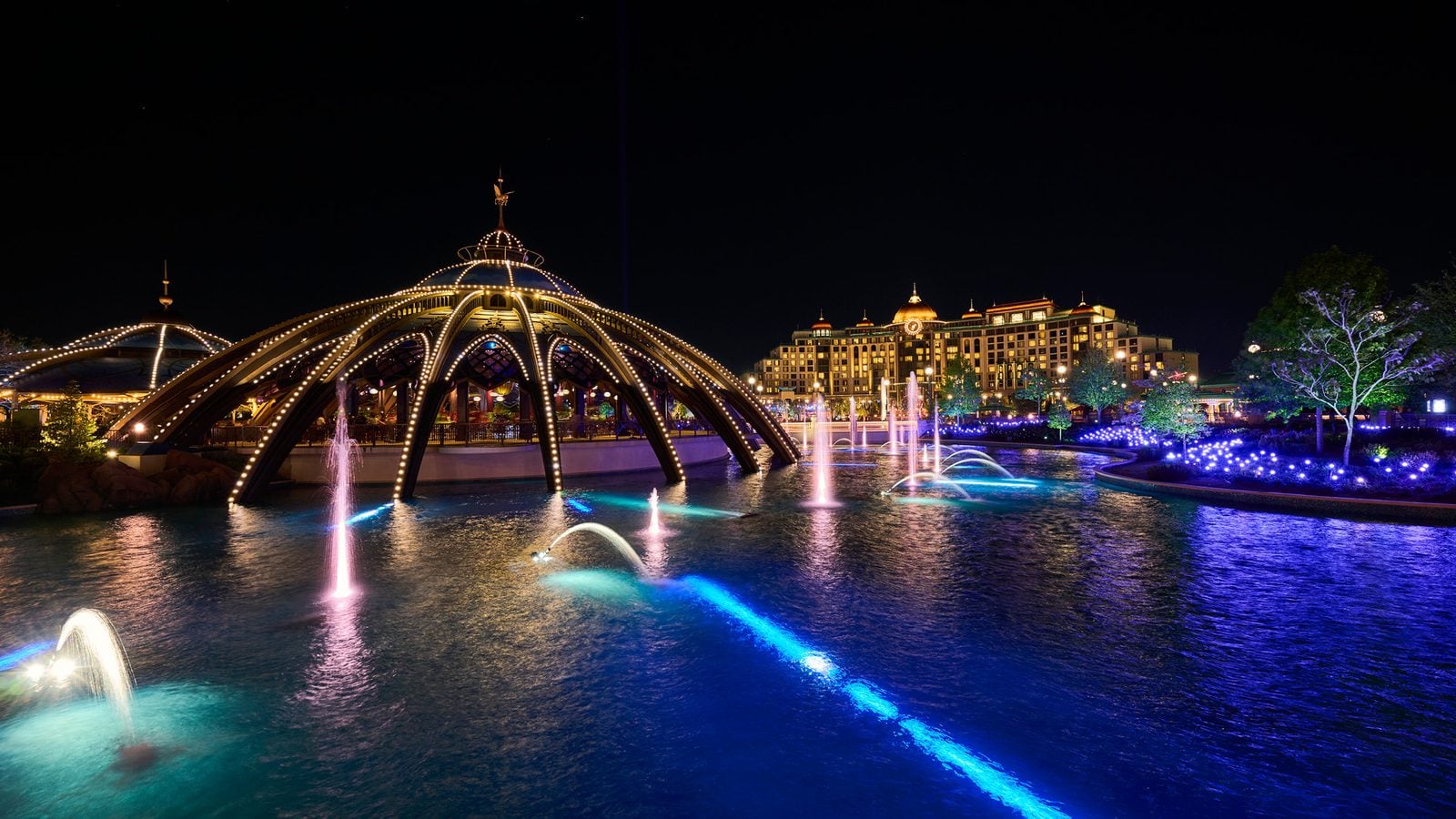 Night scene of an illuminated arched pavilion over a fountain lit lake with a lit hotelcomplex in the background