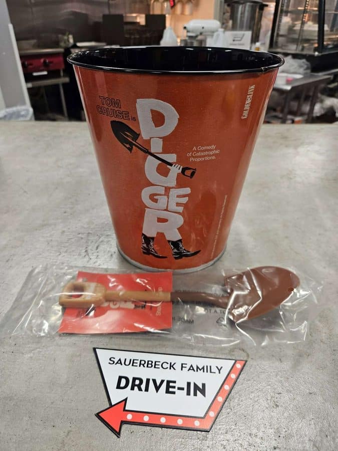 Large orange disposable cup with white graphics and a rocket illustration on a counter a SAUERBECK FAMILY DRIVE IN sign is on the floor in front