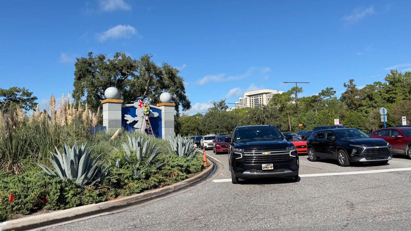 Parking lot with a landscaped island of tall grasses and agave a decorative entrance pillar with globes and a worker on a ladder near a mural under a clear blue sky