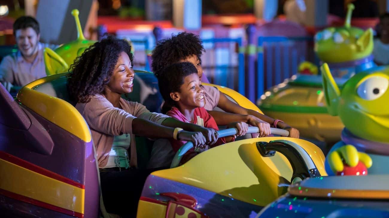 Three children smiling as they ride in a colorful bumper car at an amusement park attraction with bright yellow and purple cars nearby