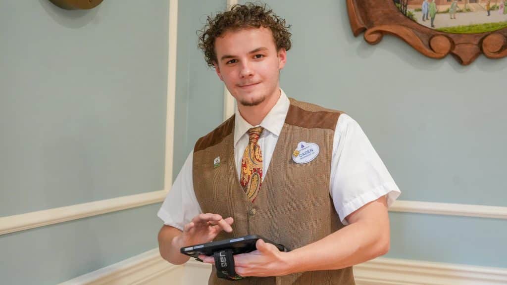 Young man in a brown plaid vest and tie uses a tablet in a pastel colored room with a decorative frame nearby informative portrait