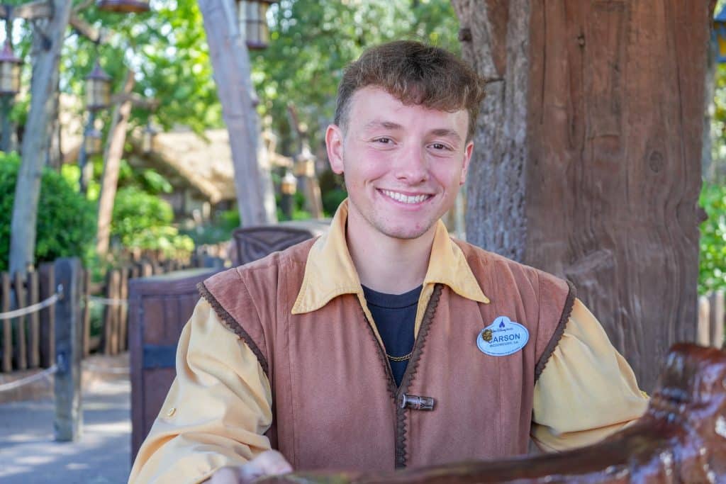 Smiling young man in a brown frontier vest and yellow sleeves poses outdoors wearing a name badge on his chest at a rustic park setting