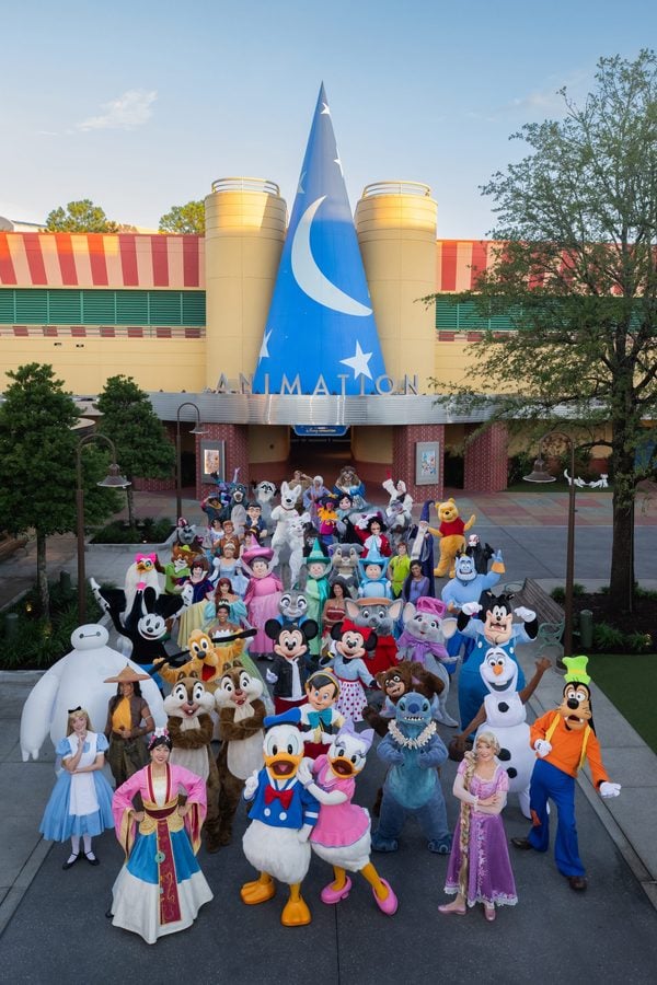 Group of costumed Disney style characters and people posing outdoors in front of a colorful animation studio building